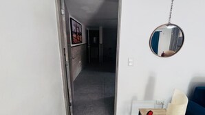 Un corridoio con una porta che conduce a una stanza, uno specchio a parete e un piccolo tavolo con una lampada e un oggetto rosso.
