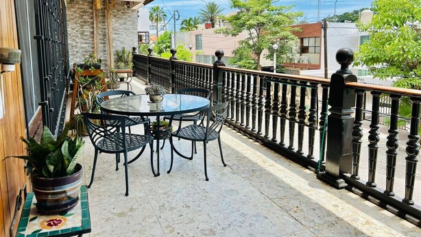 Een balkon met een glazen tafel en stoelen, een zwarte metalen reling en potplanten.