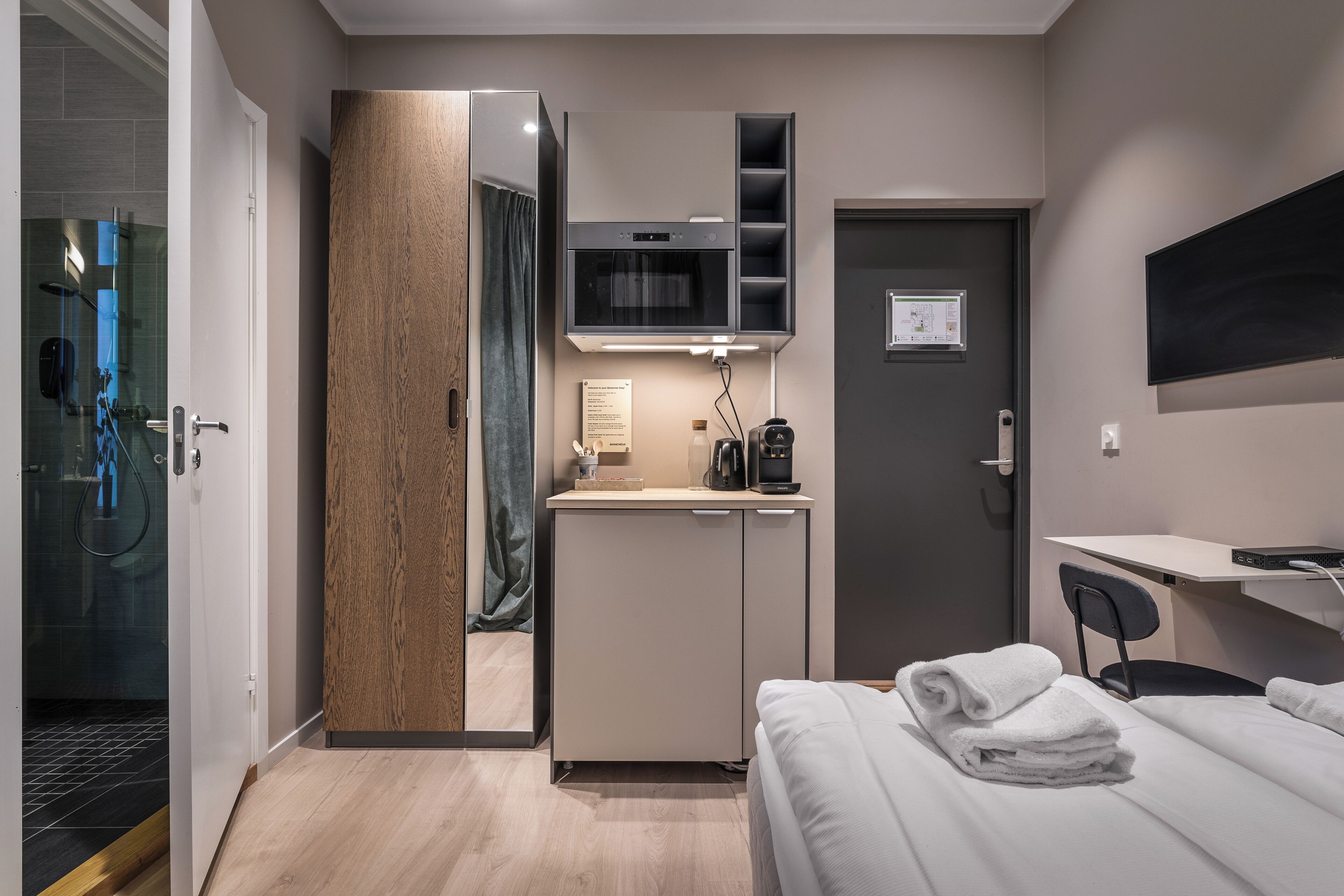 A modern hotel room with a bed, a desk, a microwave, a small kitchenette, and a shower area.
