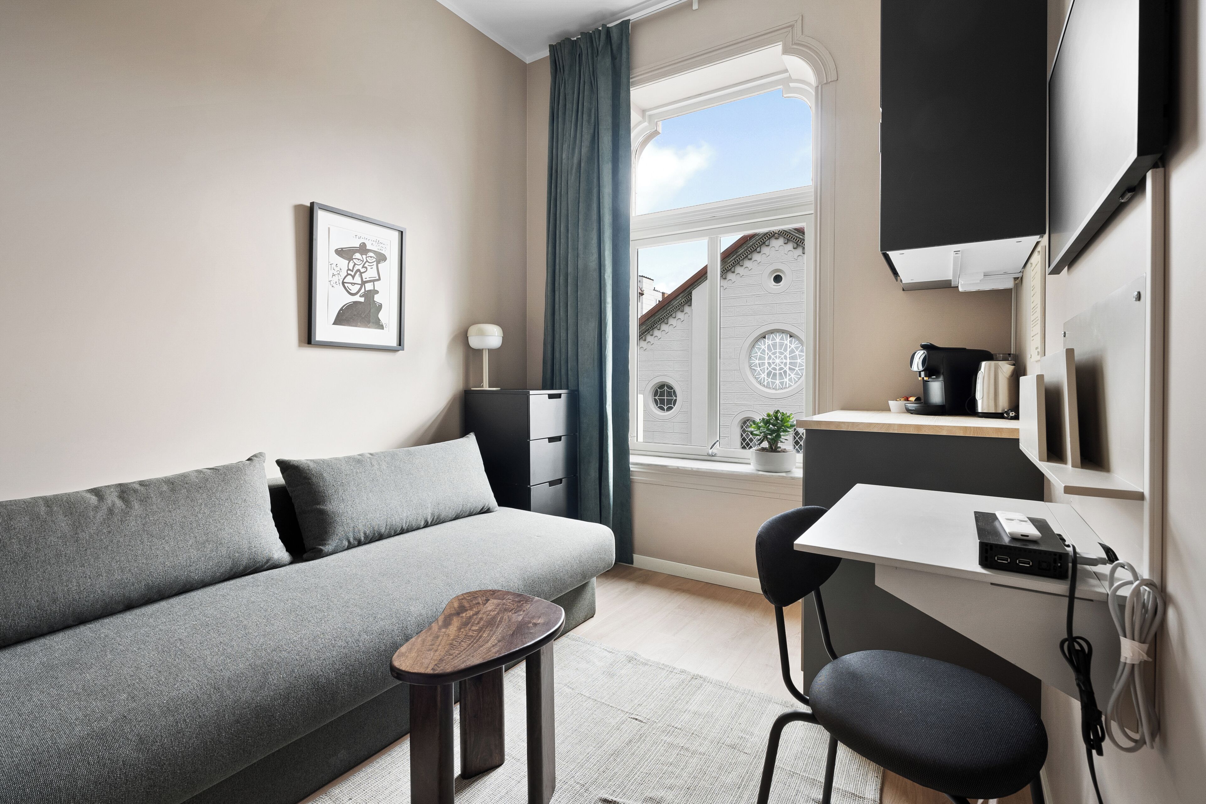 Una habitación moderna con un sofá gris, una pequeña mesa de madera, una silla negra, un escritorio con una computadora y un distribuidor de energía, una ventana con vistas a un edificio y un cuadro en la pared.