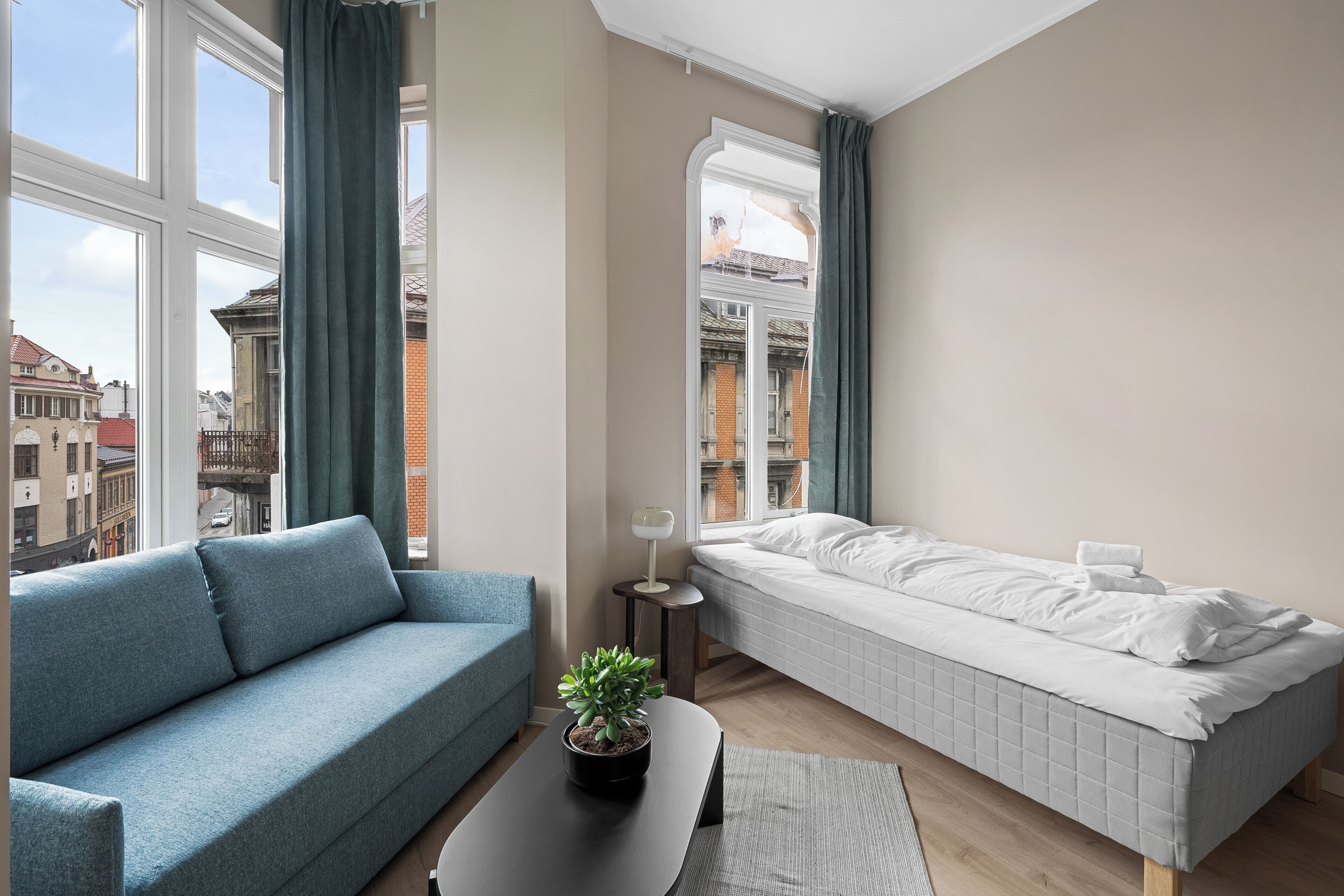 Una stanza moderna con un divano, un letto, un tavolino con una pianta e vista sui palazzi attraverso ampie finestre.