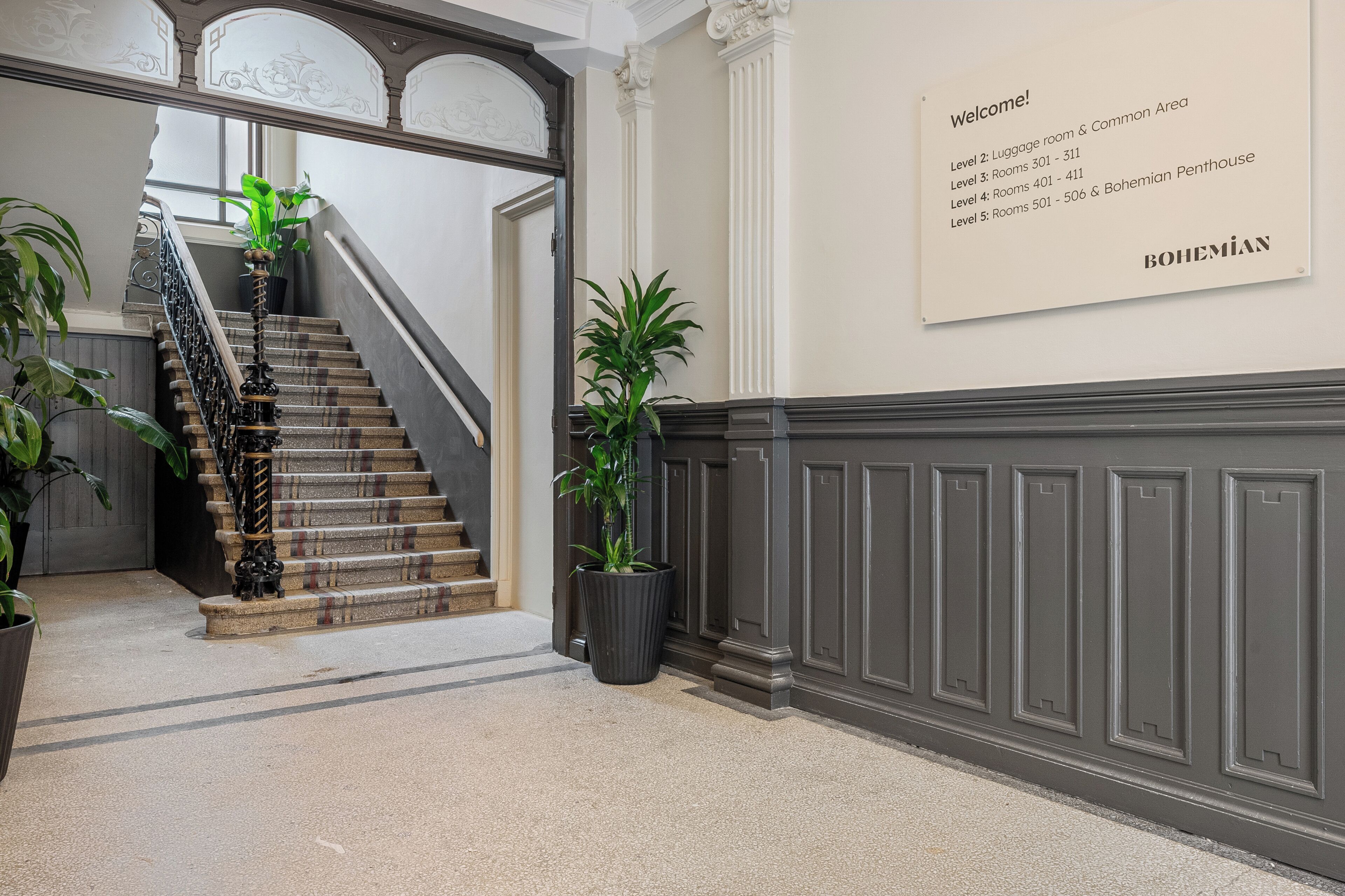 A hotel lobby with a staircase, potted plants, and a welcome sign listing room numbers.