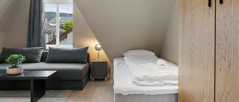 A modern loft bedroom with a sloped ceiling, a bed, a sofa, a nightstand, and a wooden wardrobe.