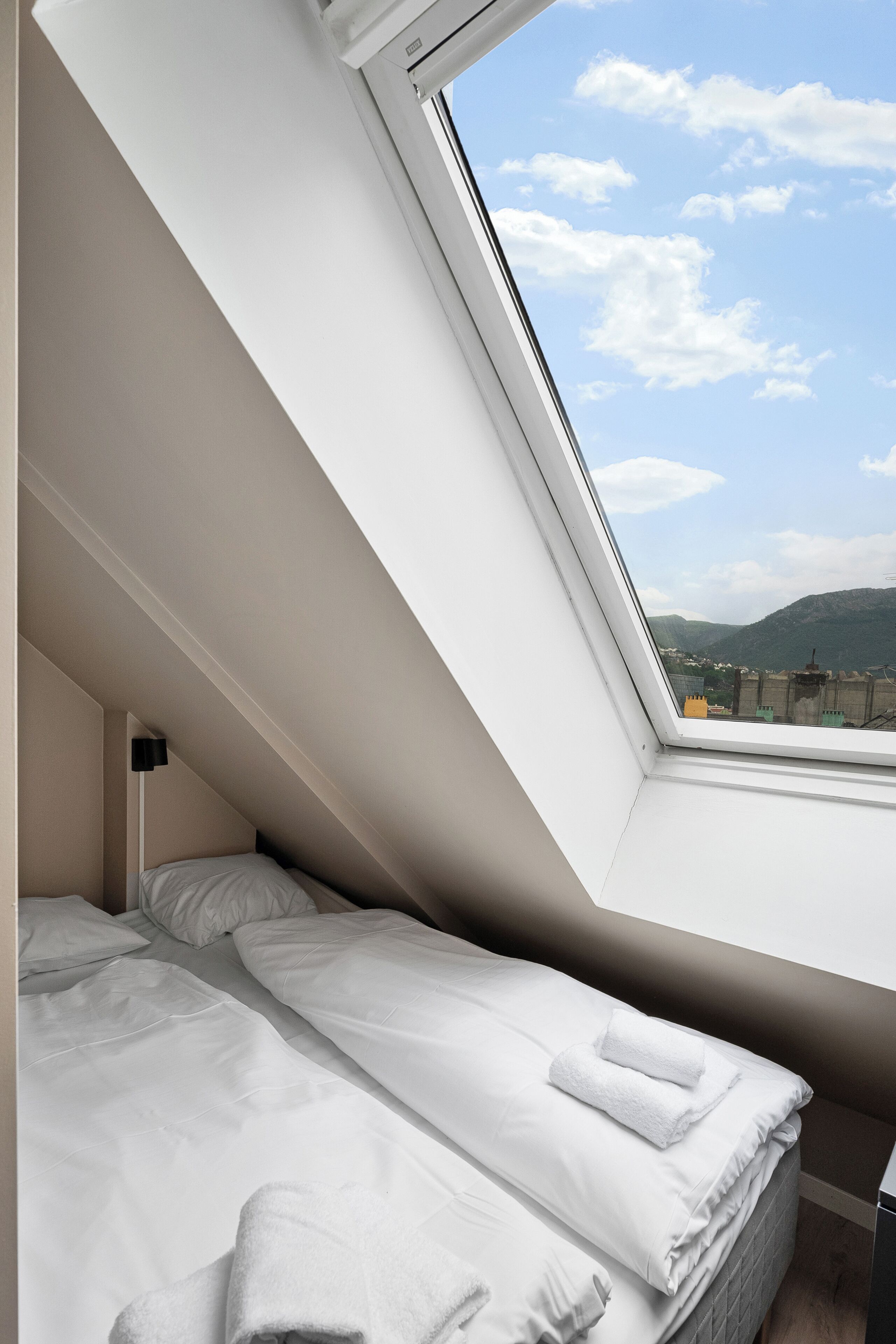 A bedroom with a skylight, white bedding, and a view of a cityscape and mountains.