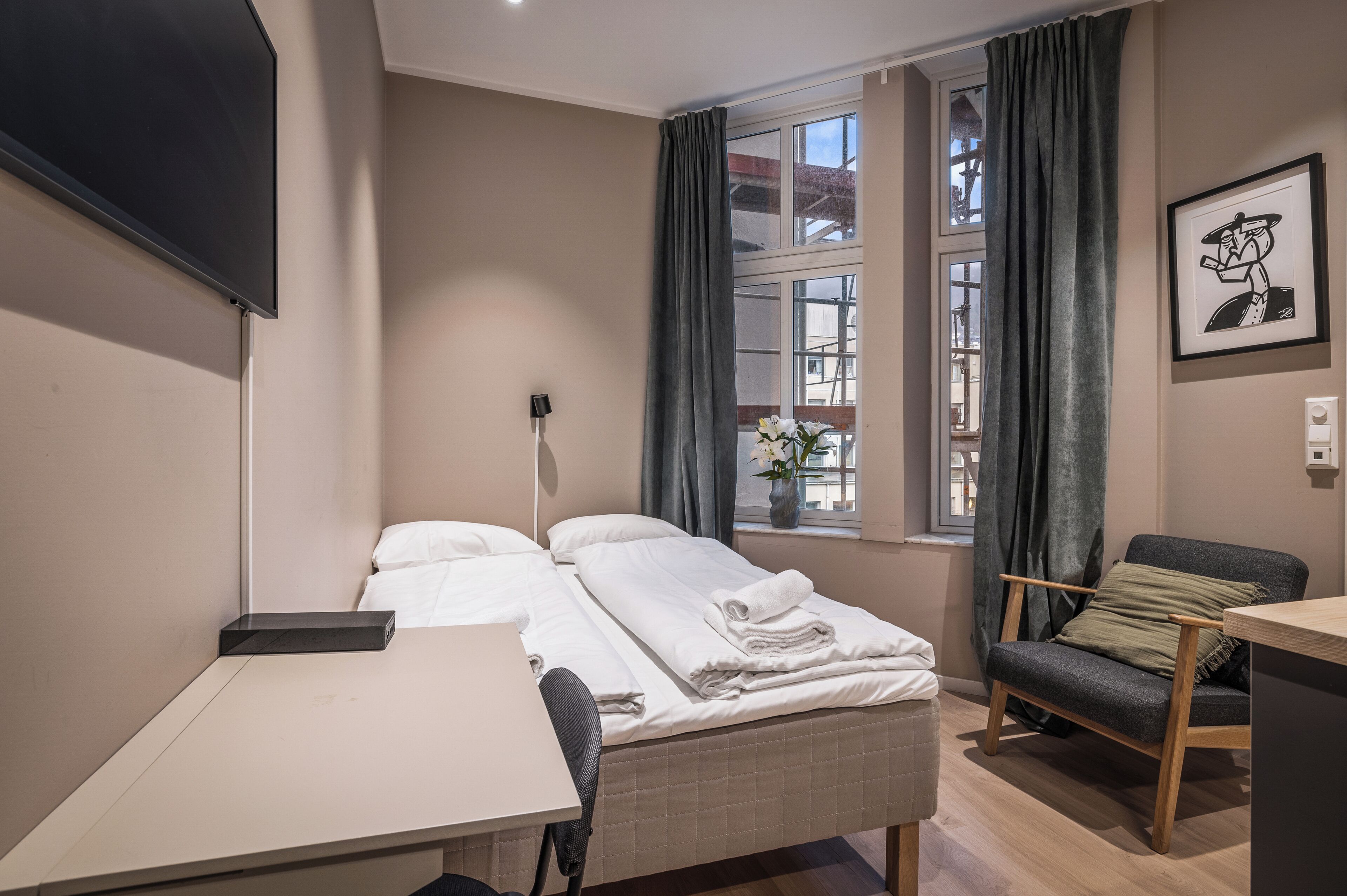 Une chambre d’hôtel avec un lit, un bureau, une chaise, une fenêtre avec des rideaux et un tableau encadré au mur.