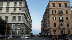Una vista di strada con due grandi edifici ai lati, cielo sereno e alcuni passanti.