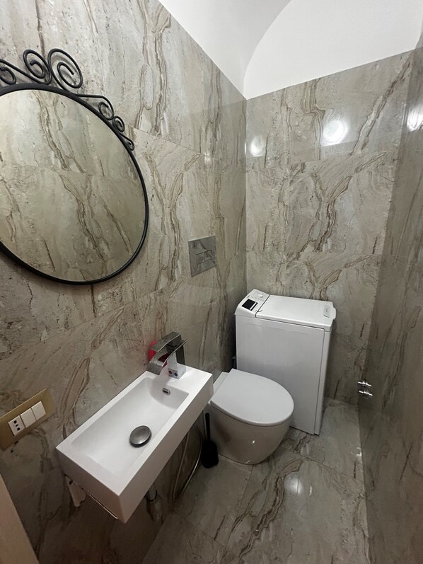Un baño con pared de mármol, un lavamanos blanco, un inodoro y un espejo con marco decorativo.