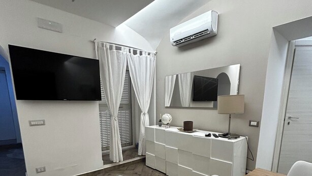 Una stanza moderna e luminosa, con televisione a schermo piatto, un armadio bianco, un tavolo da pranzo in legno e un tappeto grigio e soffice.