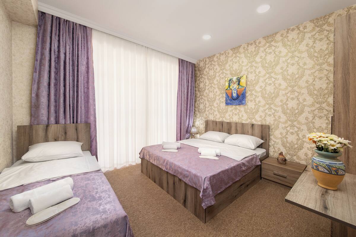 Habitación de hotel con dos camas, cabecero de madera, mesita de noche, un jarrón con flores y un cuadro en la pared.