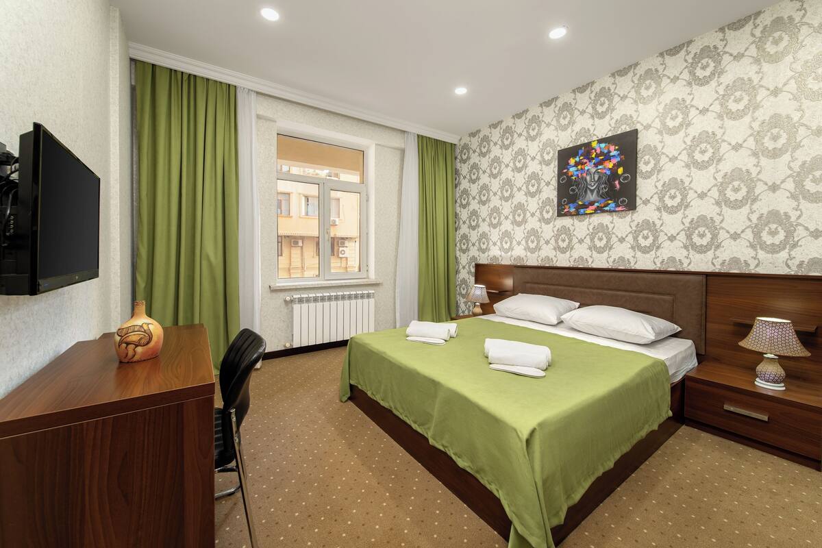 Habitación de hotel con cama, escritorio, silla, televisor, ventana con cortinas, radiador, lámpara y un cuadro en la pared.