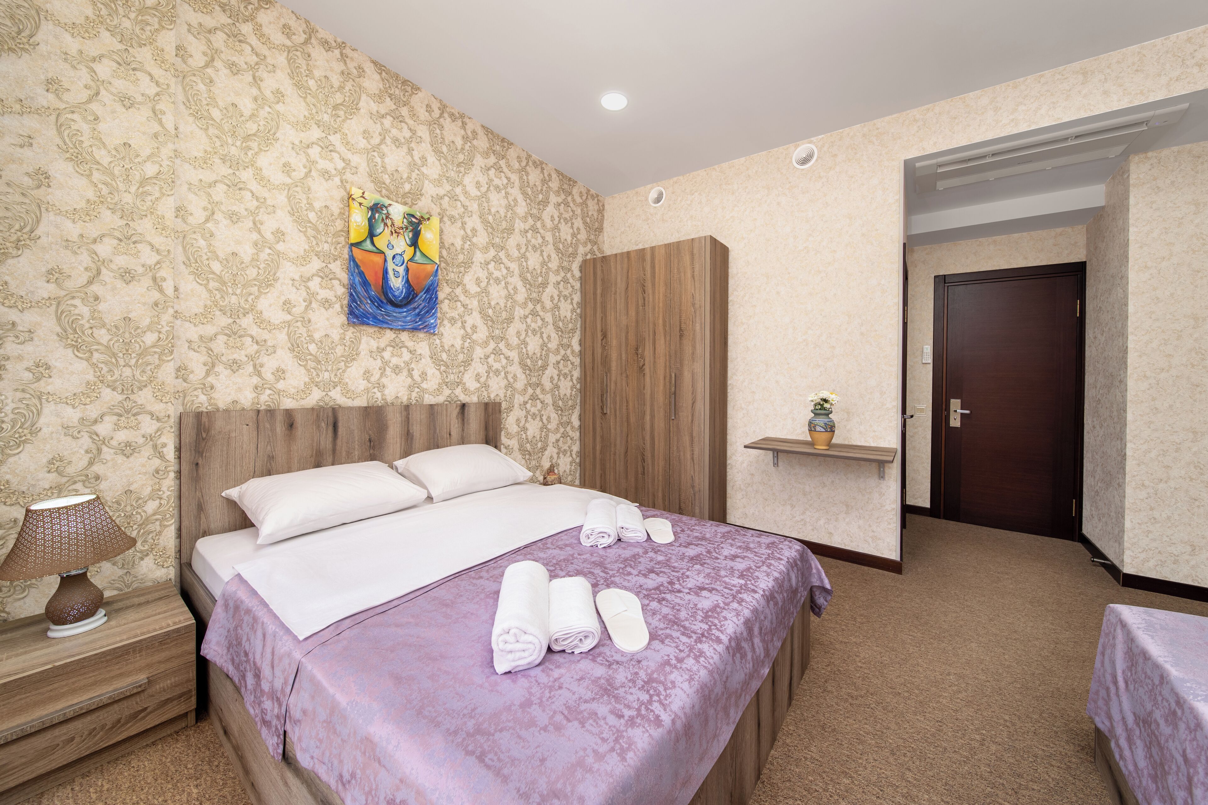 Ein Hotelzimmer mit Bett, Nachttisch mit Lampe, Holzschrank, kleiner Ablagefläche mit Pflanze und Tür.