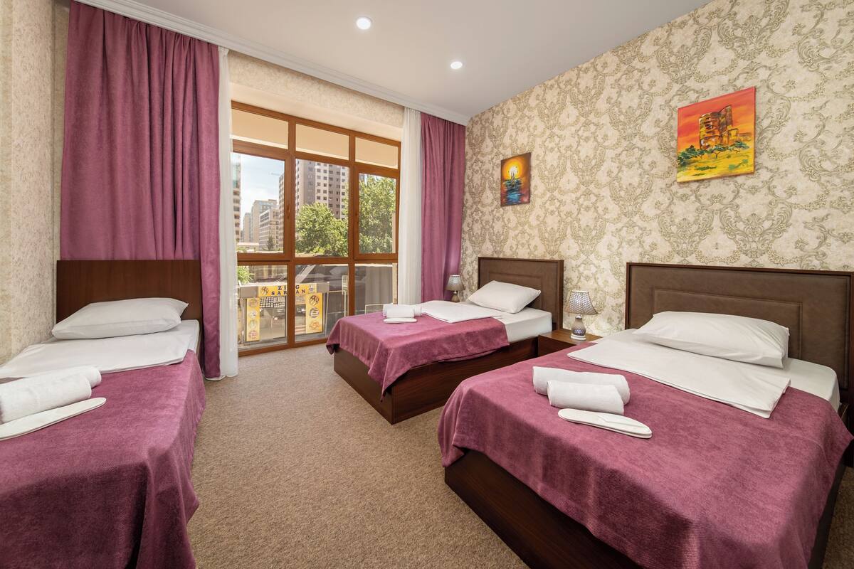 Habitación de hotel con dos camas, cada una con una colcha morada y almohadas blancas. Se ve una ventana con cortinas, un cuadro en la pared y vistas a edificios.