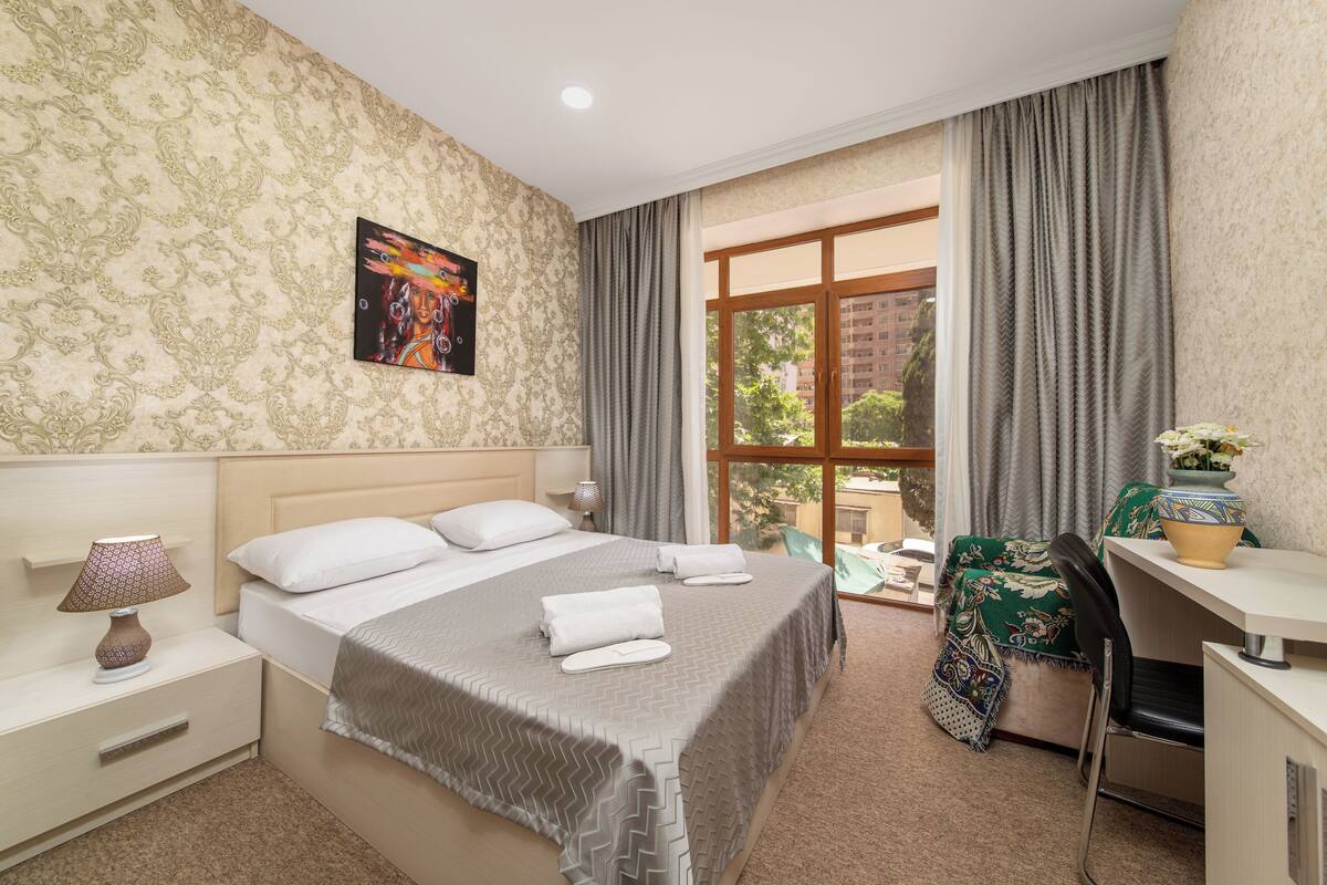 Habitación de hotel con cama, escritorio, silla, lámpara y ventana con cortinas.