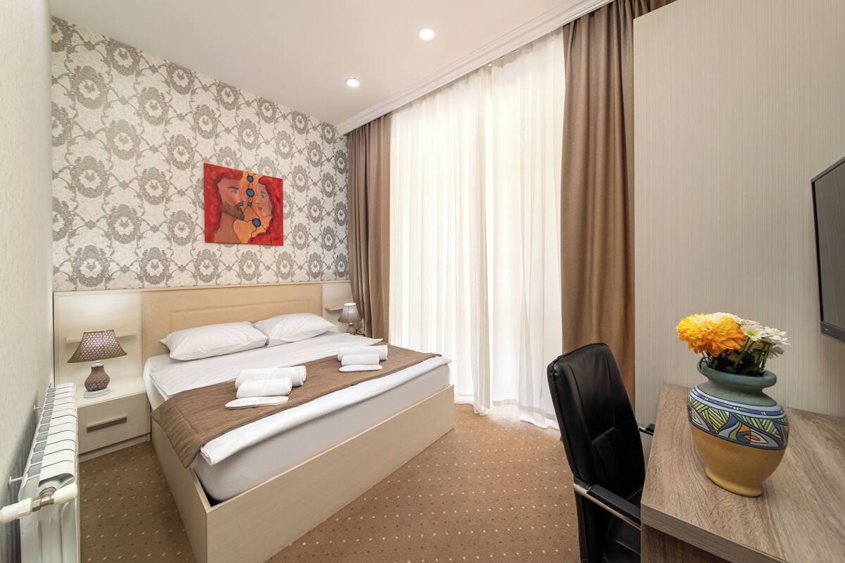 Una habitación de hotel con una cama, una silla, un escritorio, un florero con flores y un cuadro en la pared.