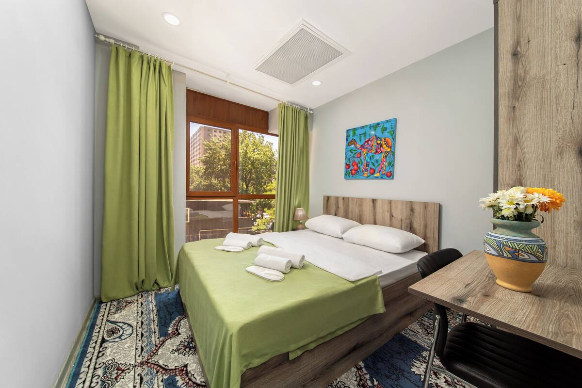 Habitación de hotel con cama, cortinas verdes, una ventana, un cuadro y un florero con flores.