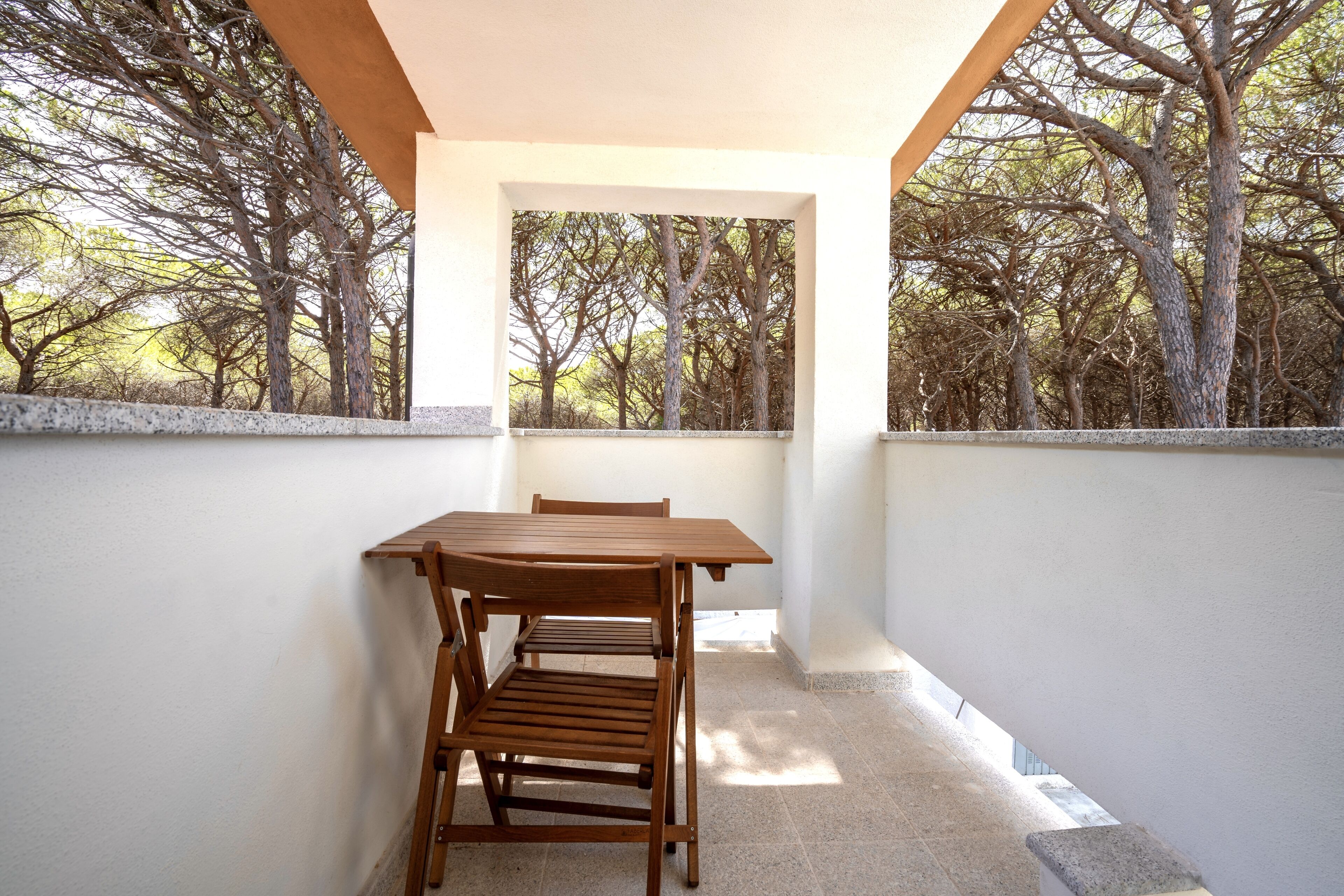 A balcony with a wooden table and chairs, white walls, and a view of trees.