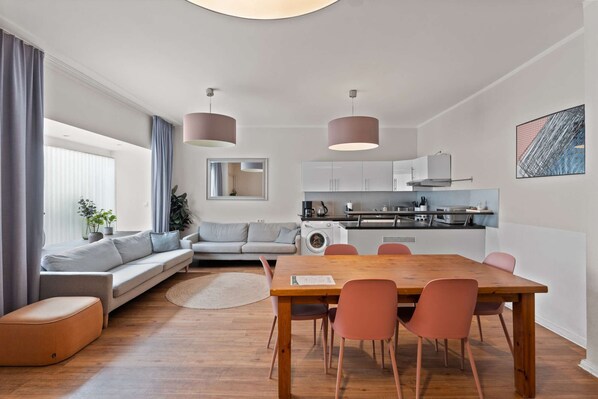 A modern apartment with a dining area, sofa, kitchen, and a large window with curtains.