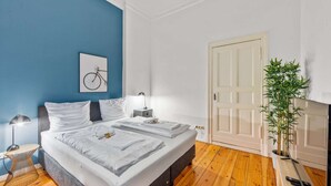 Habitación de hotel con cama doble, mesita de noche con lámpara, cuadro en la pared, puerta y planta en maceta.