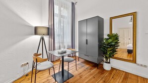 Una habitación compacta con suelo de madera, un armario gris, una mesita con una lámpara, una silla y un espejo grande.