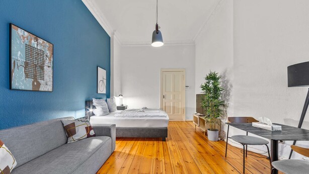 Una habitación moderna con suelo de madera, un sofá gris, una cama, un escritorio con una silla y una planta en maceta.