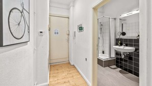 A hallway leading to a bathroom with a shower, a sink, and a mirror. There is a door at the end of the hallway and a framed picture on the wall. - primeflats - Apartment Leberstr 58 Nr. 3 (Berlin)