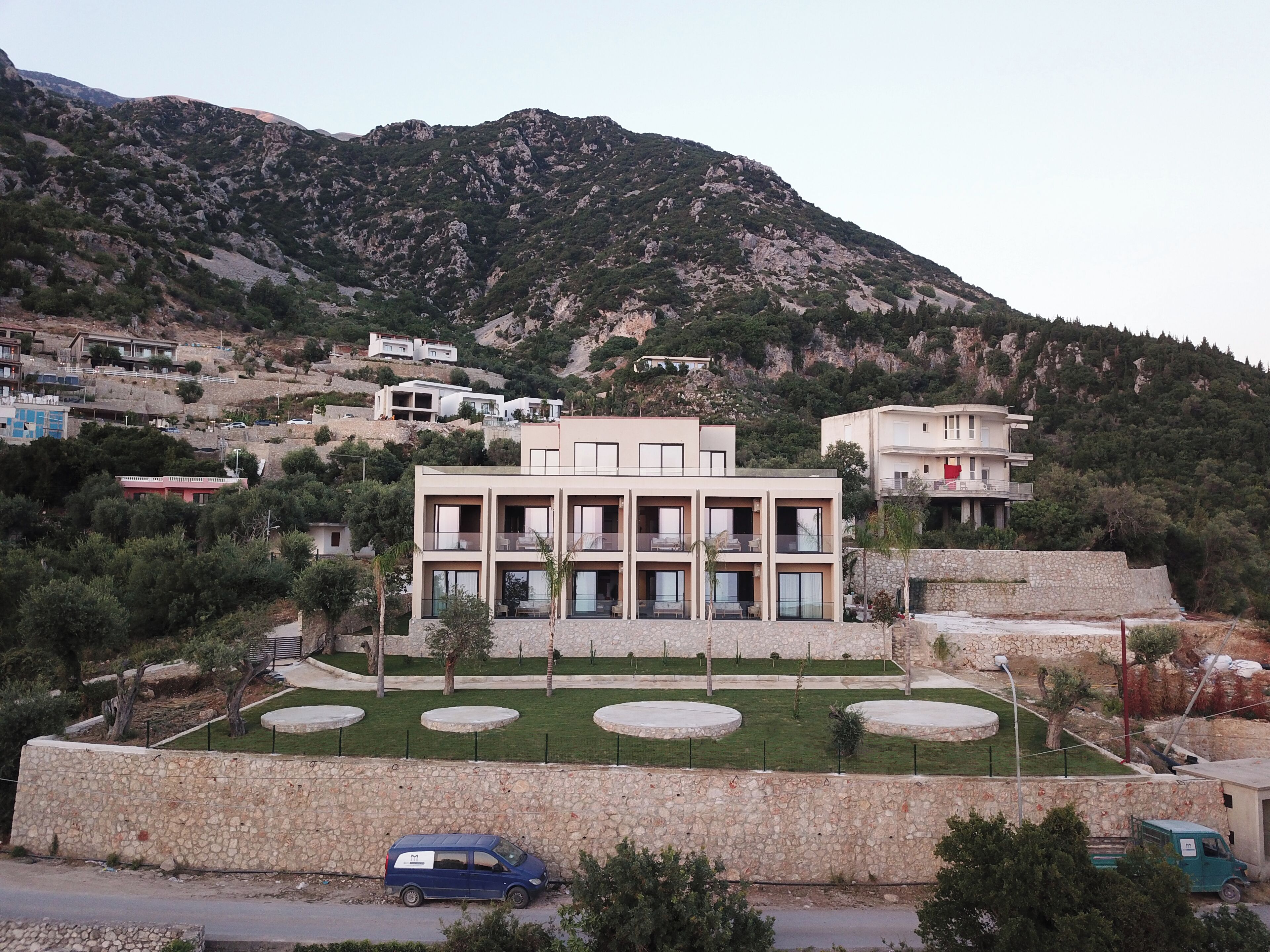 A modern multi-story building with a stone facade, surrounded by a landscaped garden with circular planters, set against a mountainous backdrop.