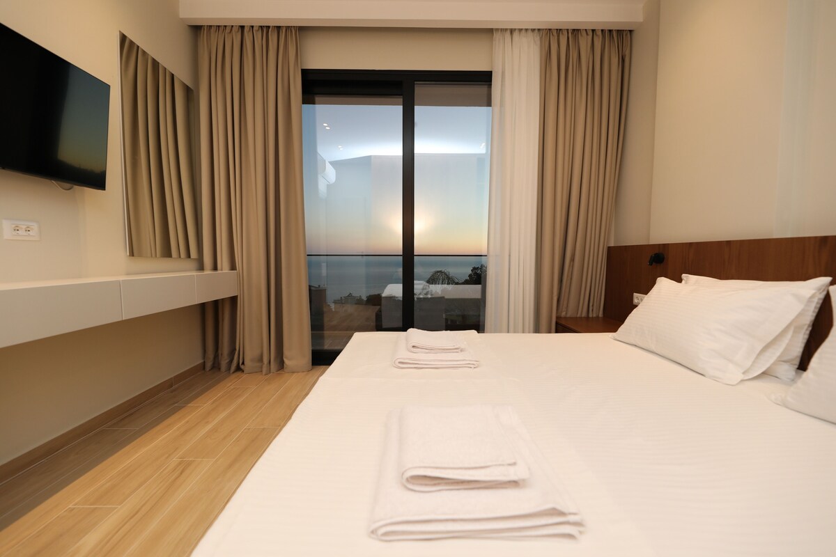 A hotel room with a large bed, a flat-screen TV mounted on the wall, and a view through the window.
