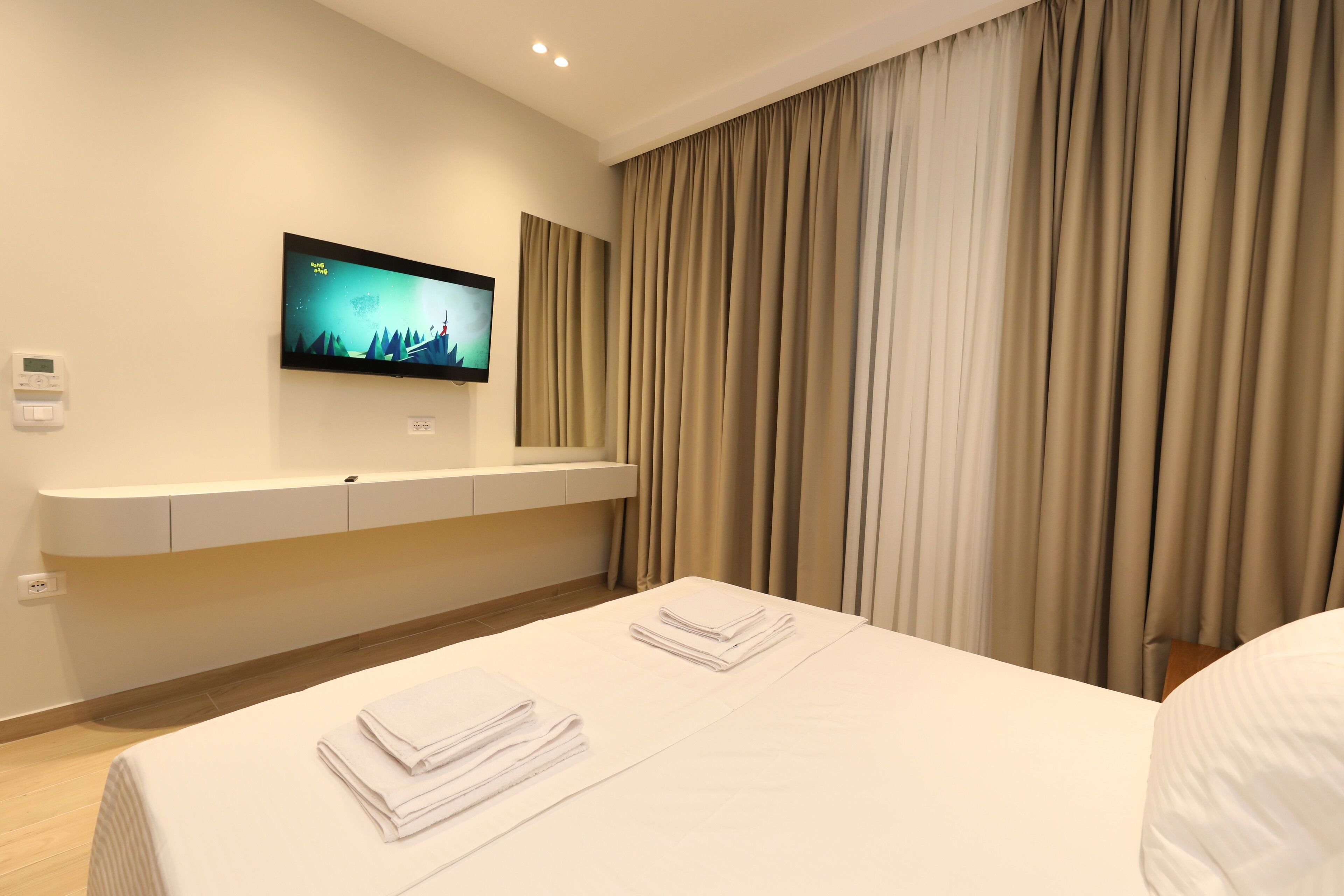 A hotel room with a bed, a flat-screen TV mounted on the wall, a bedside table, and a window with curtains.