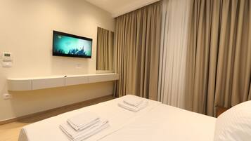 A hotel room with a bed, a flat-screen TV mounted on the wall, a bedside table, and a window with curtains.