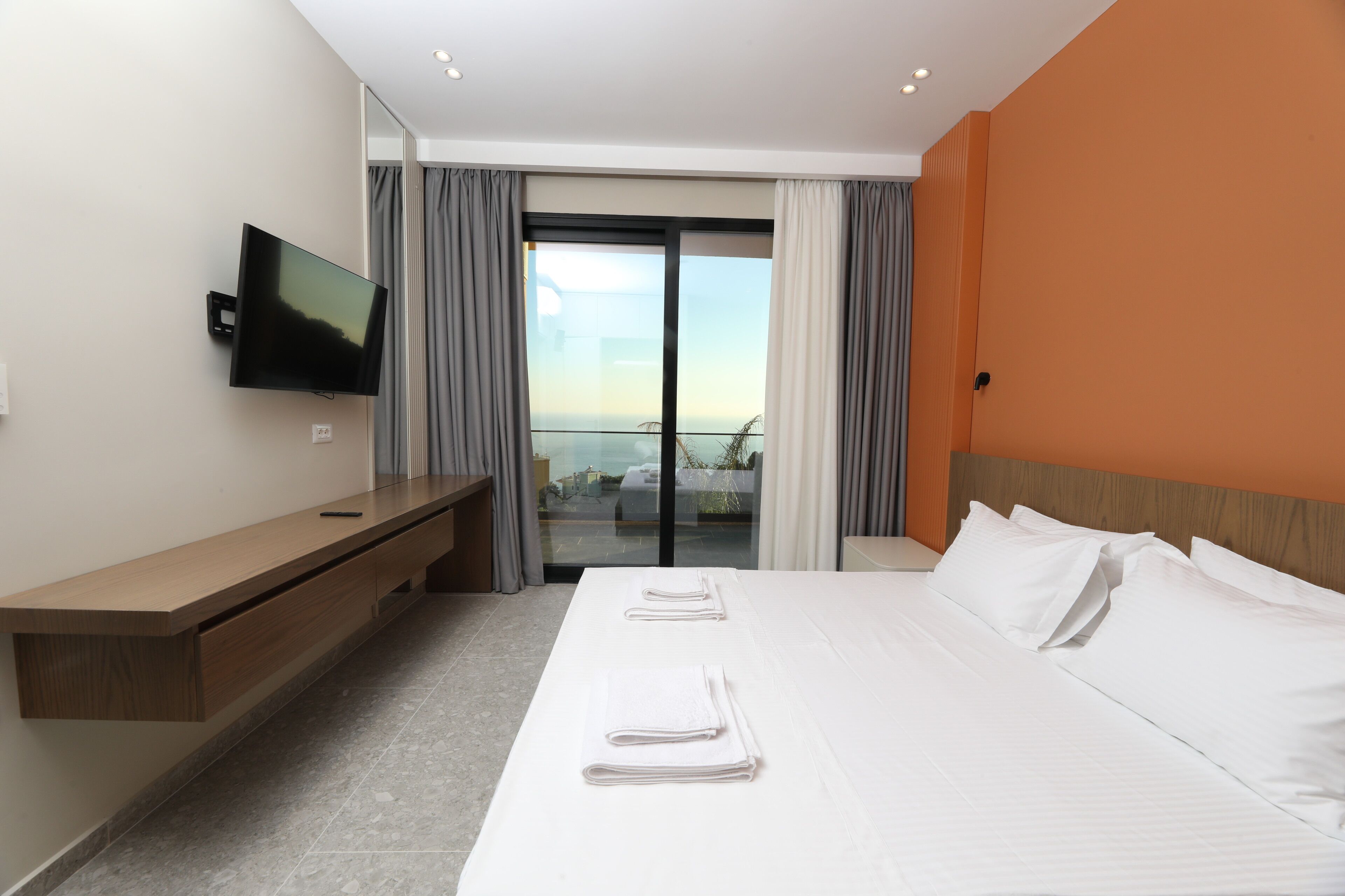 A hotel room with a bed, a TV, a desk, and a balcony with curtains.