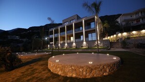 A modern hotel with multiple balconies, a stone circular patio, and a mountainous backdrop. - Aurum Hotel (Himarë)