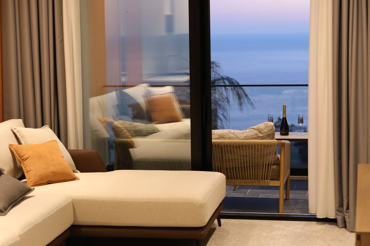 A modern living room with a sofa, a wicker chair, and a table with a bottle and glass.