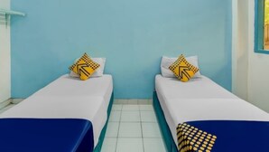 A room with two single beds, each with a white mattress and blue bedspread. There are decorative pillows with a yellow and black pattern on each bed. The walls are painted light blue, and there is a window on the right side of the image. - Hotel Margadana (Tegal)