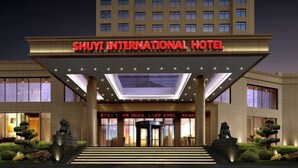 Das äußere Erscheinungsbild des Shuyi International Hotels mit einem markanten Eingang, einem überdachten Gang und einer gut beleuchteten Fassade.