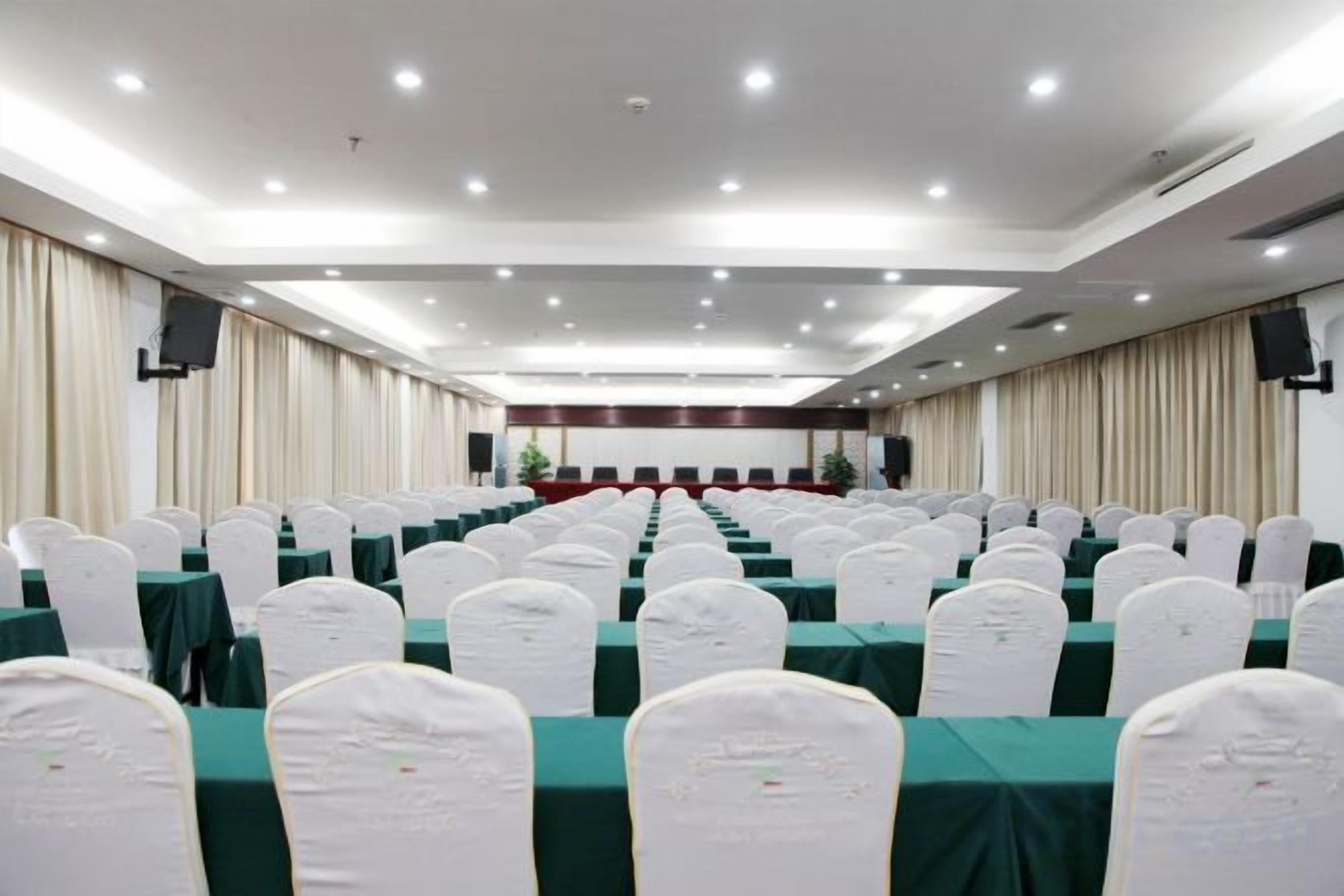 A conference room with rows of tables and chairs, a stage with a podium, and a large screen.