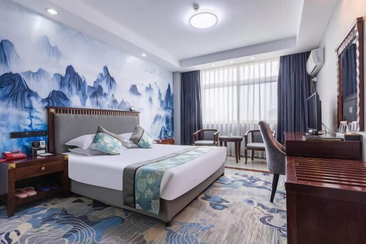 Habitación de hotel con cama, escritorio, silla, televisor y un cuadro de un paisaje montañoso.