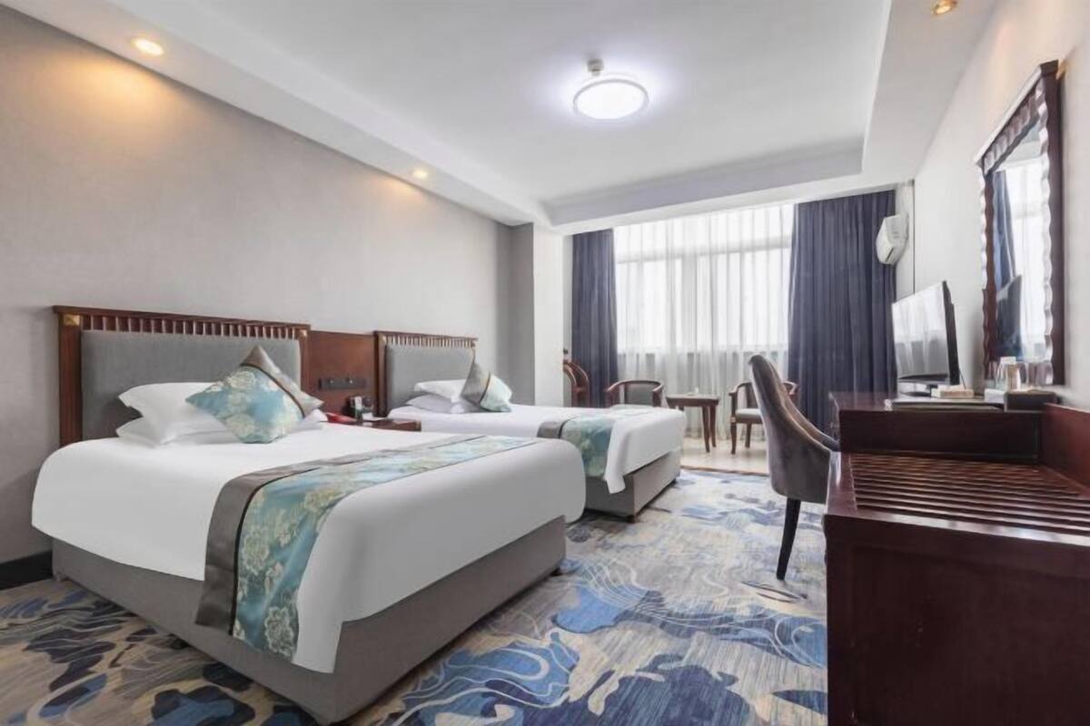 Habitación de hotel con dos camas, un escritorio, una silla, un ventanal con cortinas e iluminación en el techo.