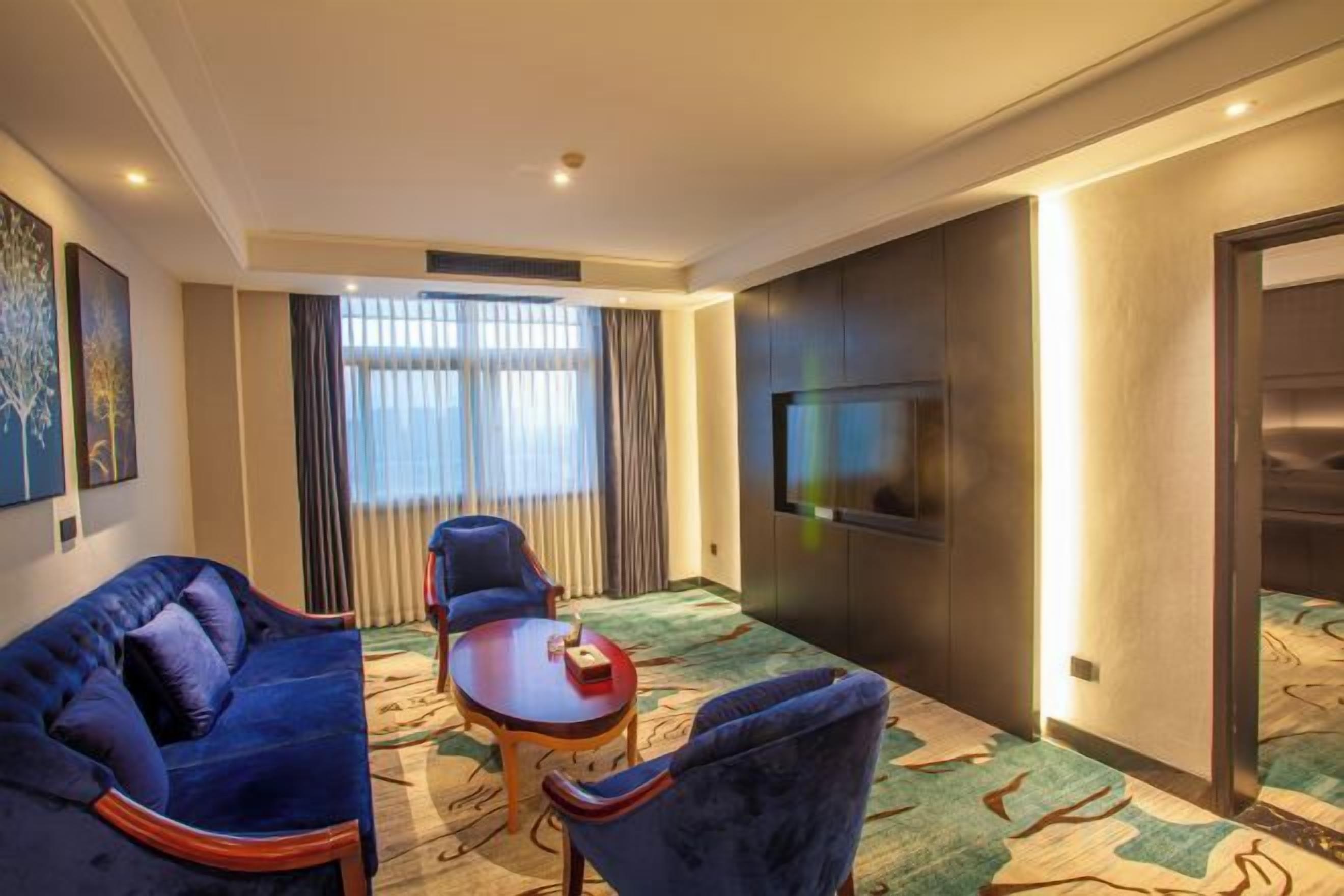 Une chambre d’hôtel avec un canapé, des fauteuils, une petite table, un téléviseur et une grande fenêtre avec des rideaux.