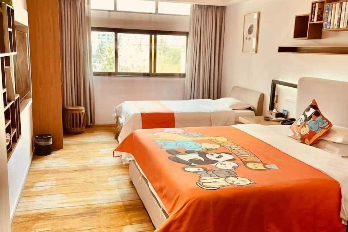 A hotel room with two beds, a wooden floor, a window with curtains, a bookshelf, and a desk with a computer.