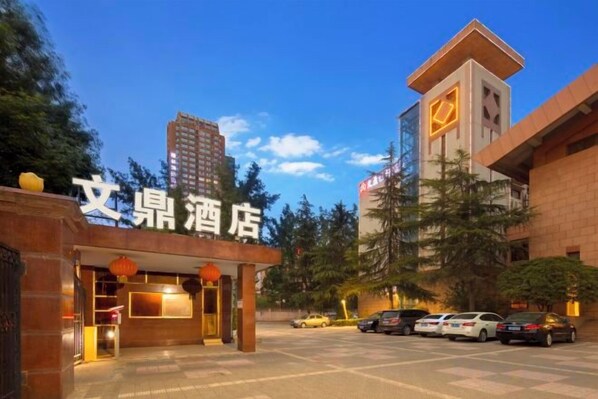 Entrada de hotel con un cartel, una zona de estacionamiento y un edificio alto al fondo.