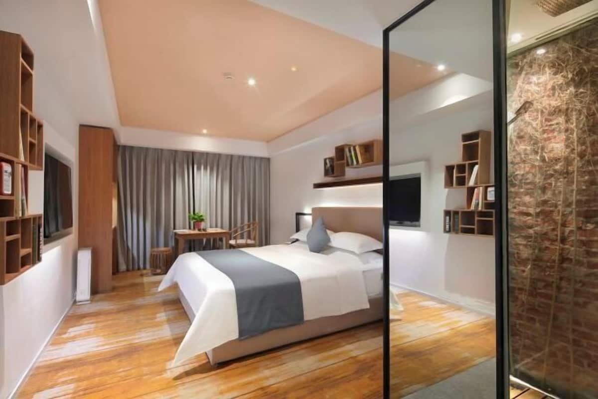 A modern bedroom with a wooden floor, a bed with a grey and white bedspread, a desk with a chair, and built-in shelving with books and decorative items.
