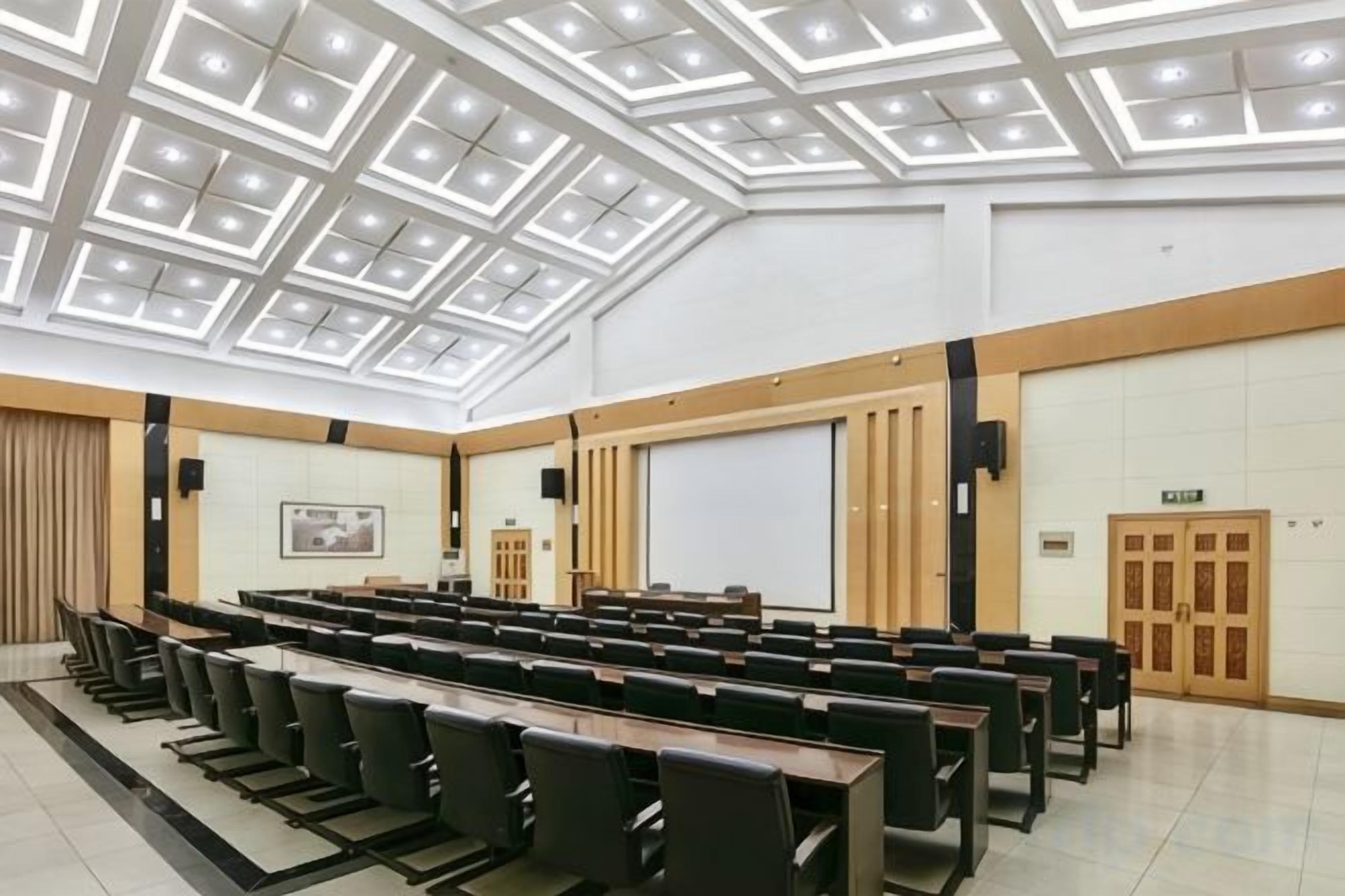 Une salle de conférence avec un plafond haut, des rangées de chaises, un écran de projection et un tableau blanc.