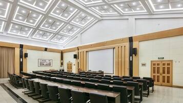 Une salle de conférence avec un plafond haut, des rangées de chaises, un écran de projection et un tableau blanc.