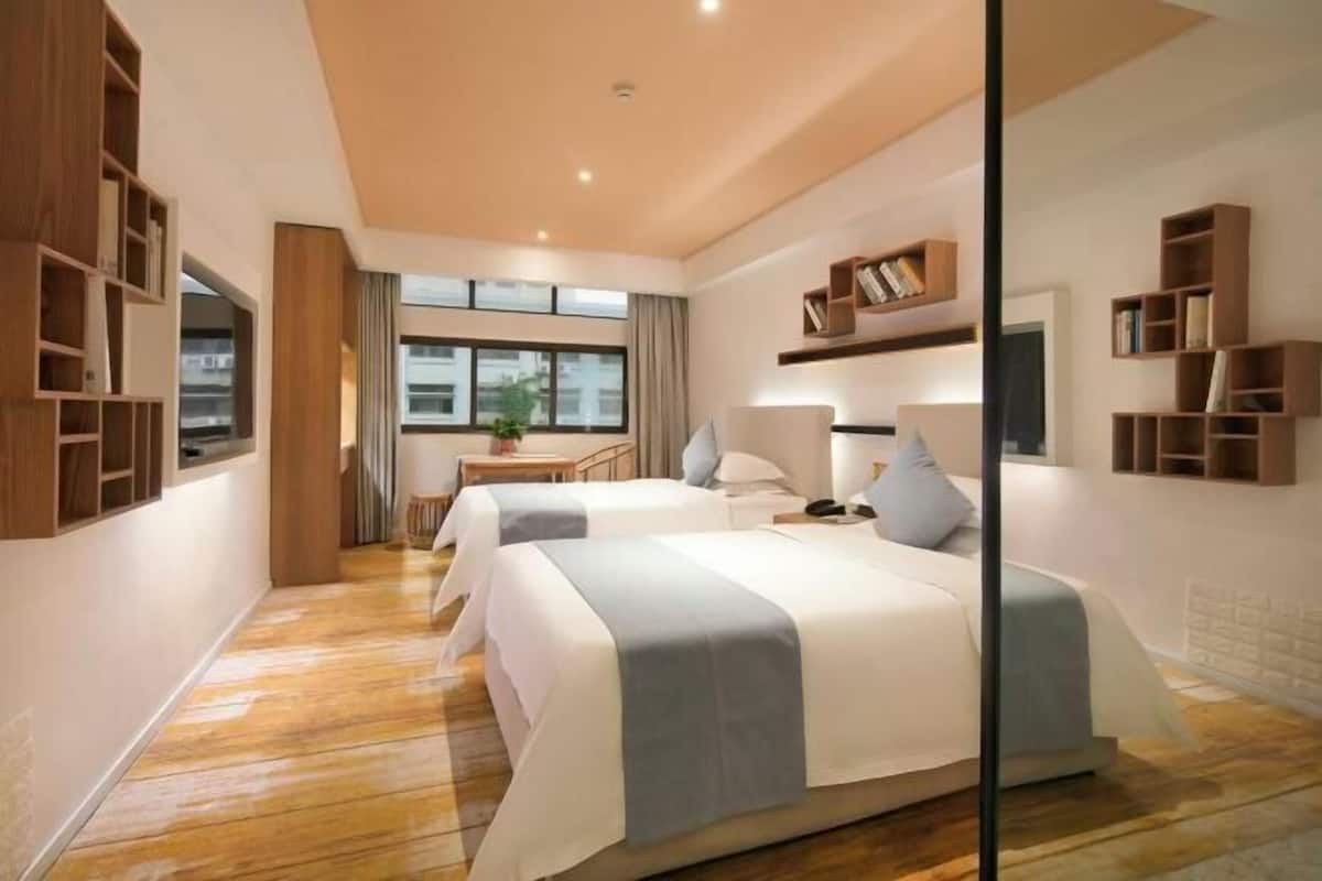 A modern hotel room with two beds, wooden shelving units, a desk with a chair, and a large window providing natural light.