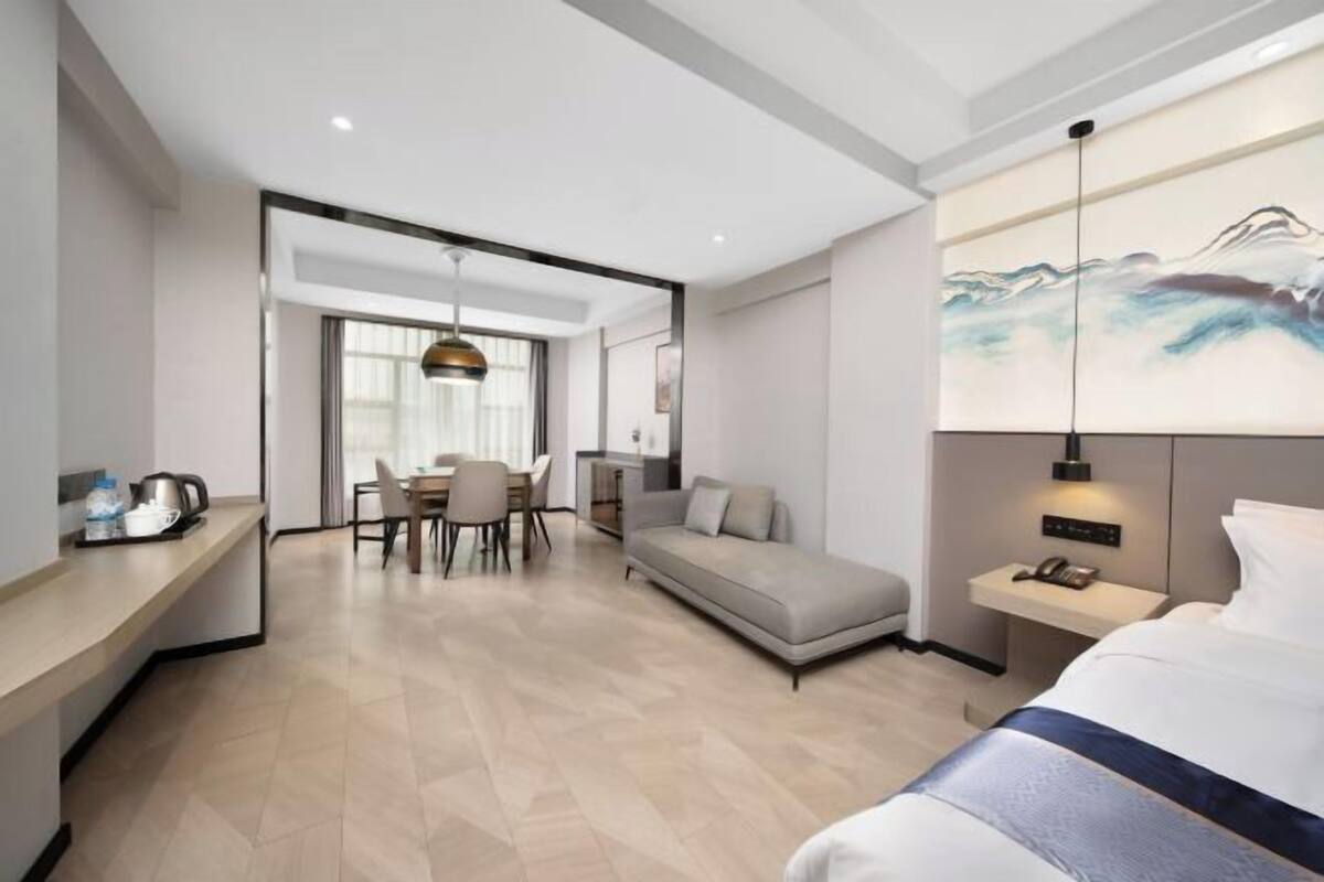 Una habitación de hotel moderna con cama, sofá, mesa de comedor con sillas, zona de cocina y un cuadro en la pared.