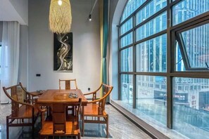 Un comedor con mesa y sillas de madera, ventanas amplias y una lámpara colgante.