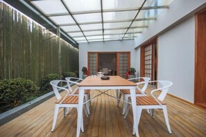Un patio techado con una mesa de madera y sillas blancas, techo de vidrio y valla de bambú.