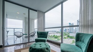 Ein geräumiges Zimmer mit großen Fenstern und Blick auf die Stadt, ein grünes, gepolstertes Sofa, ein passendes Ottomanen und zwei schwarze Stühle mit Drahtgestell.