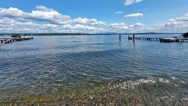 Ein ruhiger See mit klarem Wasser, eine hölzerne Anlegestelle, die ins Wasser führt, und eine ferne Uferlinie mit Bergen.