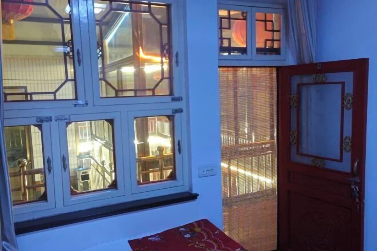 A room with a window overlooking a street, a door with decorative elements, and a red cushion on the floor.
